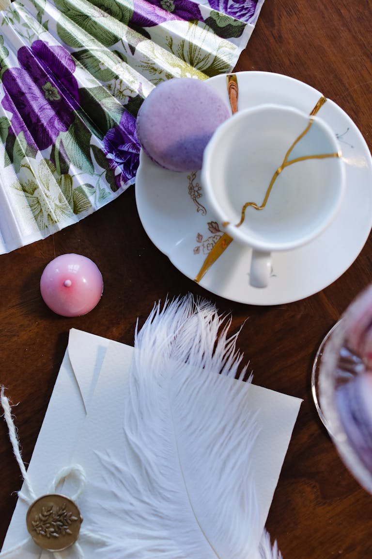 Aesthetic flatlay featuring a cup, lavender macaron, feather, and floral fan on wooden surface.